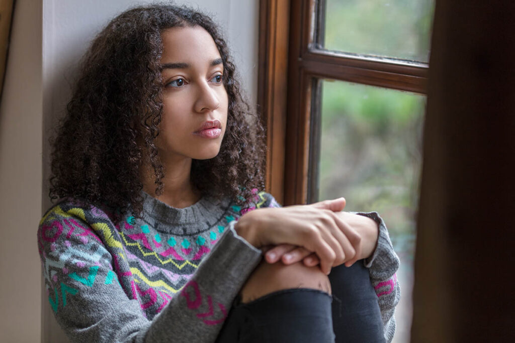 teenage girl sitting in her room by the window looking pensive and wondering if adults are skilled at recognizing self harm in teens